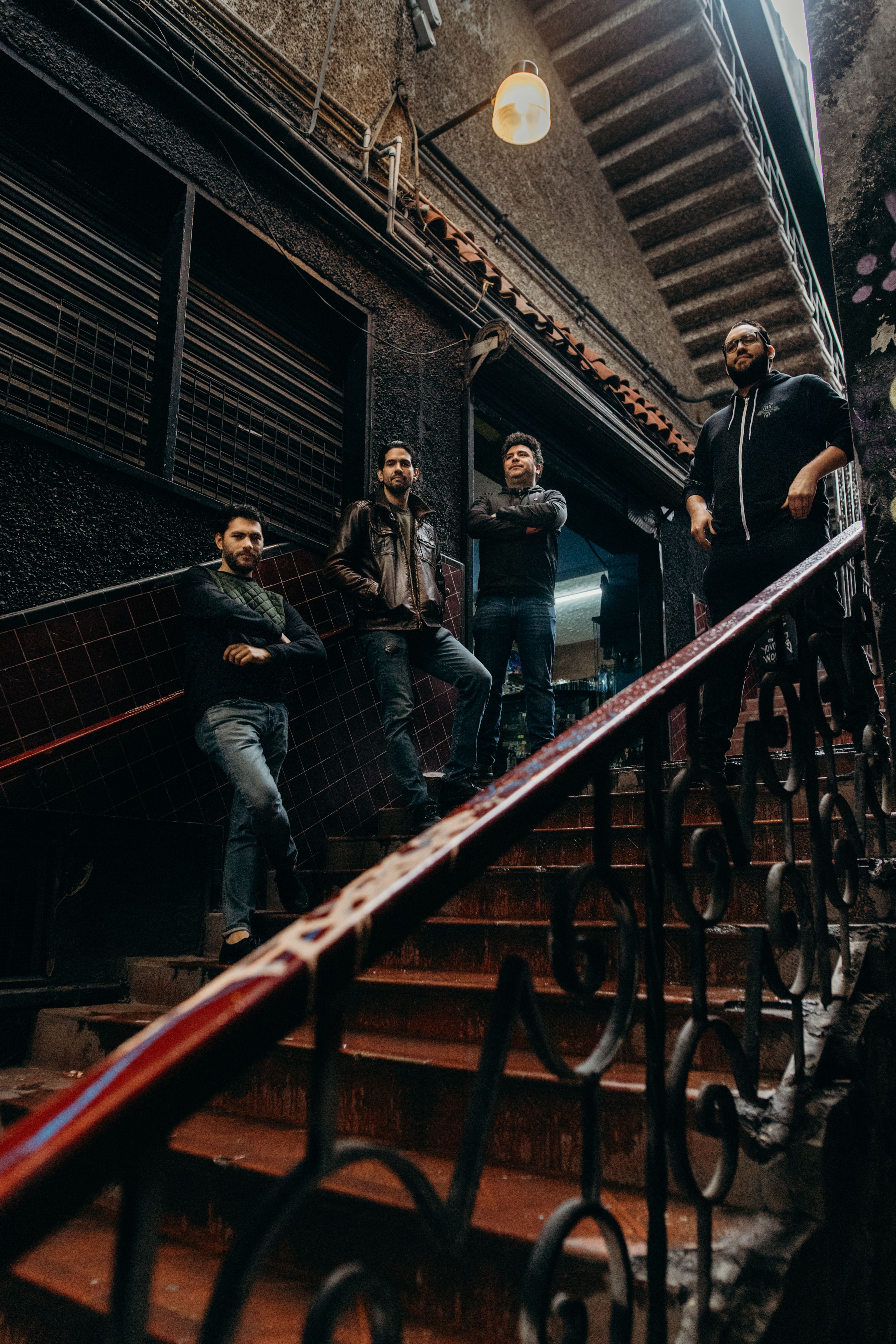 Four men standing on a dimly lit urban staircase, exuding a mysterious and edgy vibe.