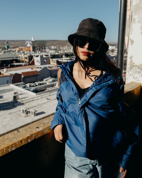 Free Woman in blue jacket and hat with sunglasses, standing on rooftop overlooking urban landscape during daytime. Stock Photo