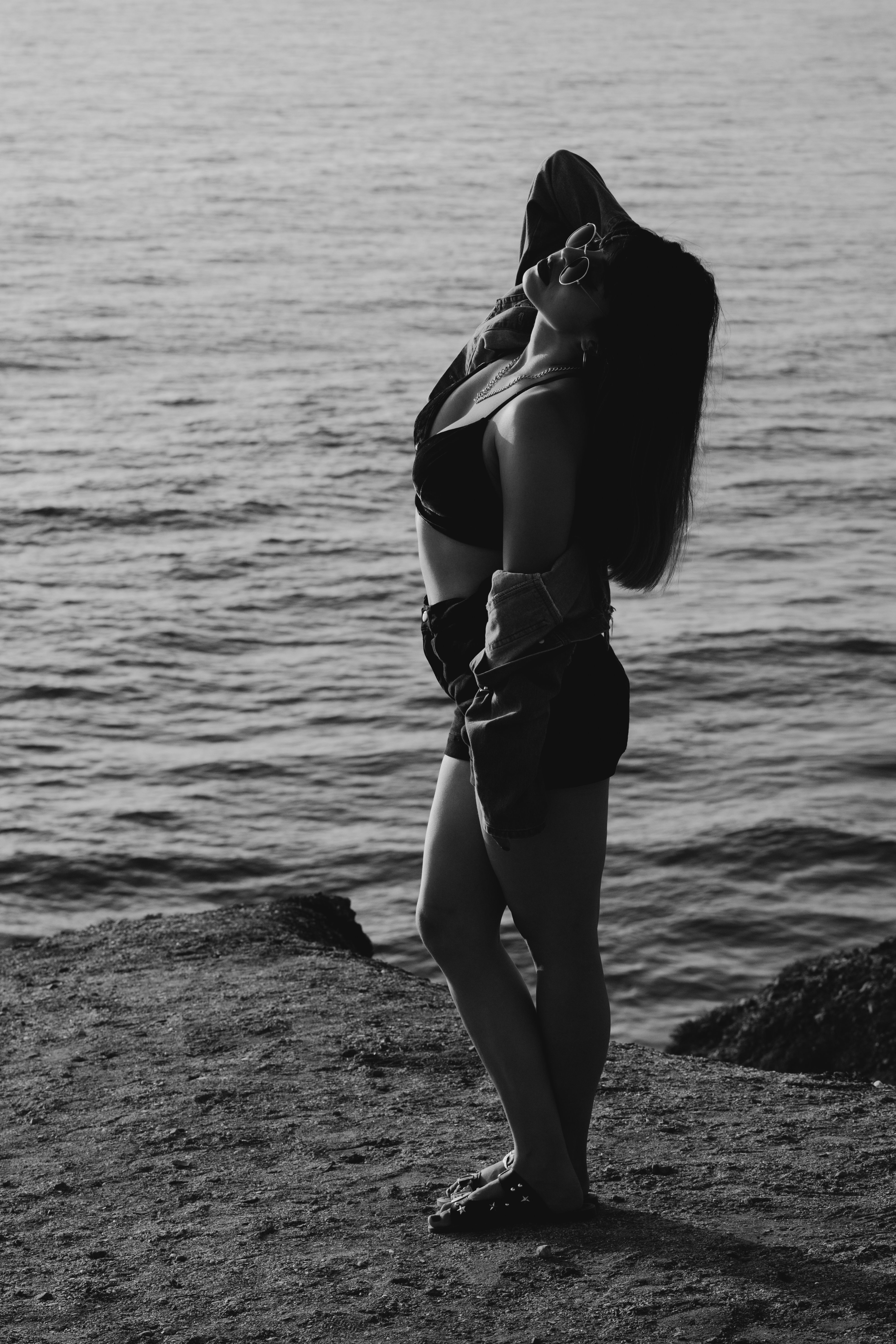 A woman poses gracefully by the sea in a black-and-white vertical shot, exuding elegance.
