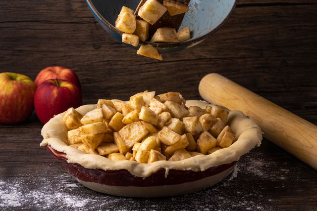 Cinnamon apple chunks tumbling from a bowl into an unbaked pie crust beside a rolling pin