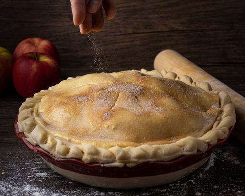 Free Close-up of a homemade apple pie being sprinkled with sugar, surrounded by apples and a rolling pin. Stock Photo