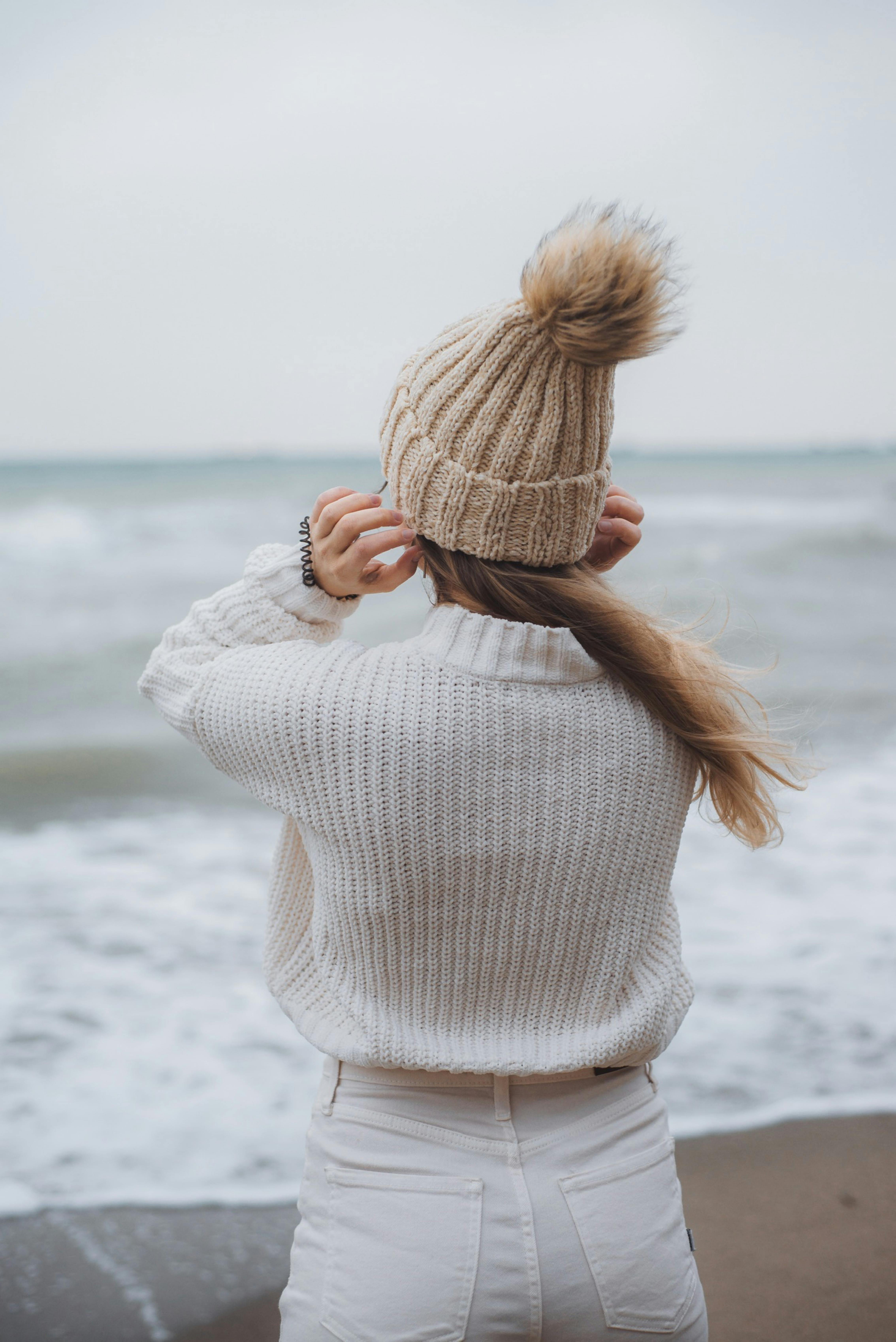 Back view of a woman in cozy winter clothing standing by the ocean, embracing the serene view.