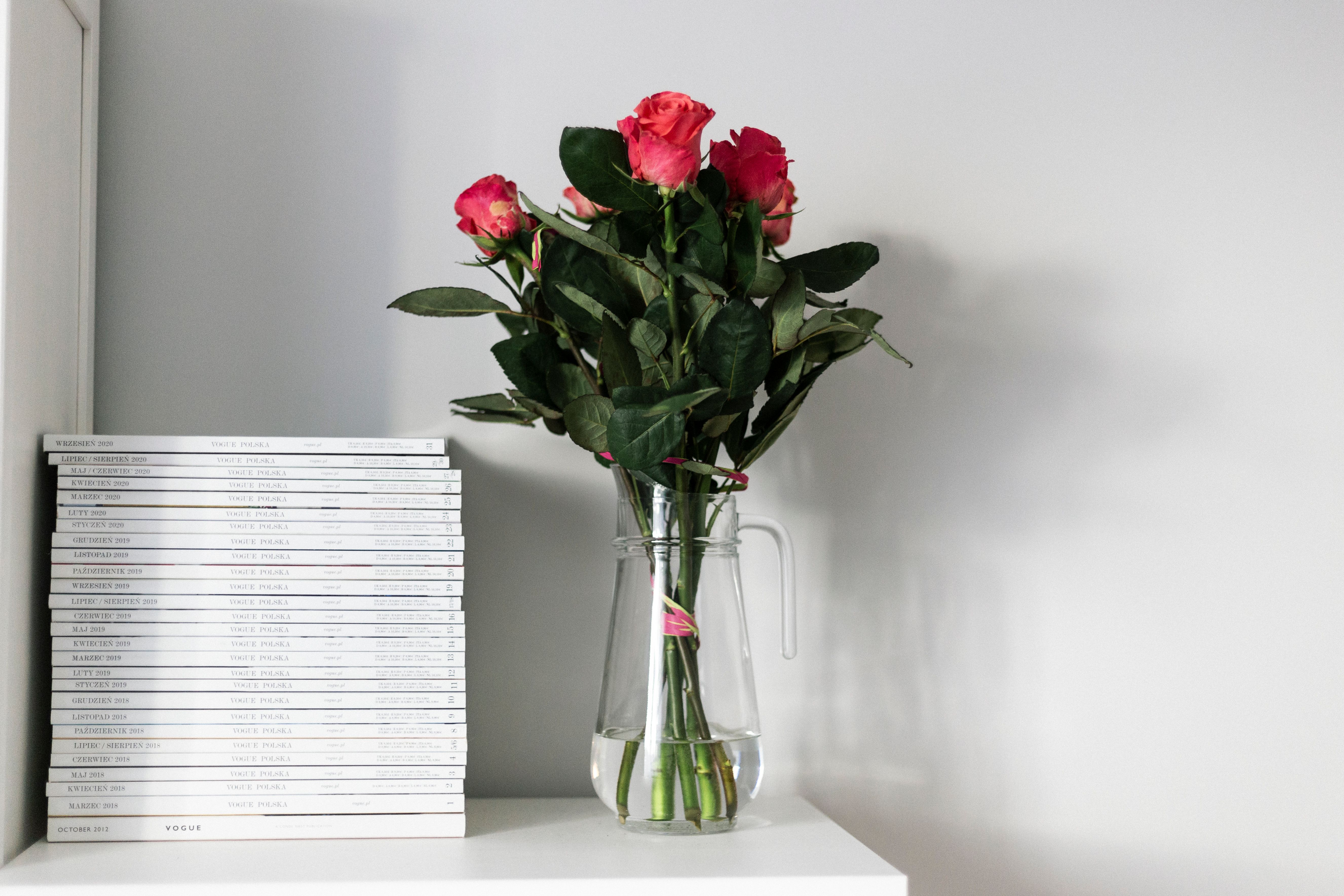 A minimalist arrangement of pink roses in a glass pitcher beside stacked magazines on a white shelf.