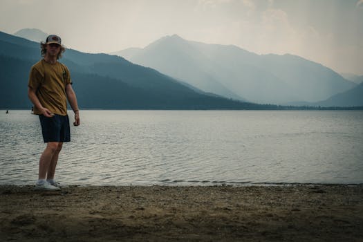 Young man standing by a serene mountain lake with fog and scenic views.