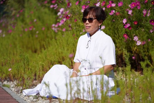 Elderly woman sitting in a flower garden wearing white clothes and sunglasses.