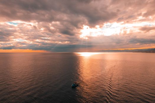 A breathtaking aerial view of a boat under dramatic clouds and sunrays over the ocean at sunset.