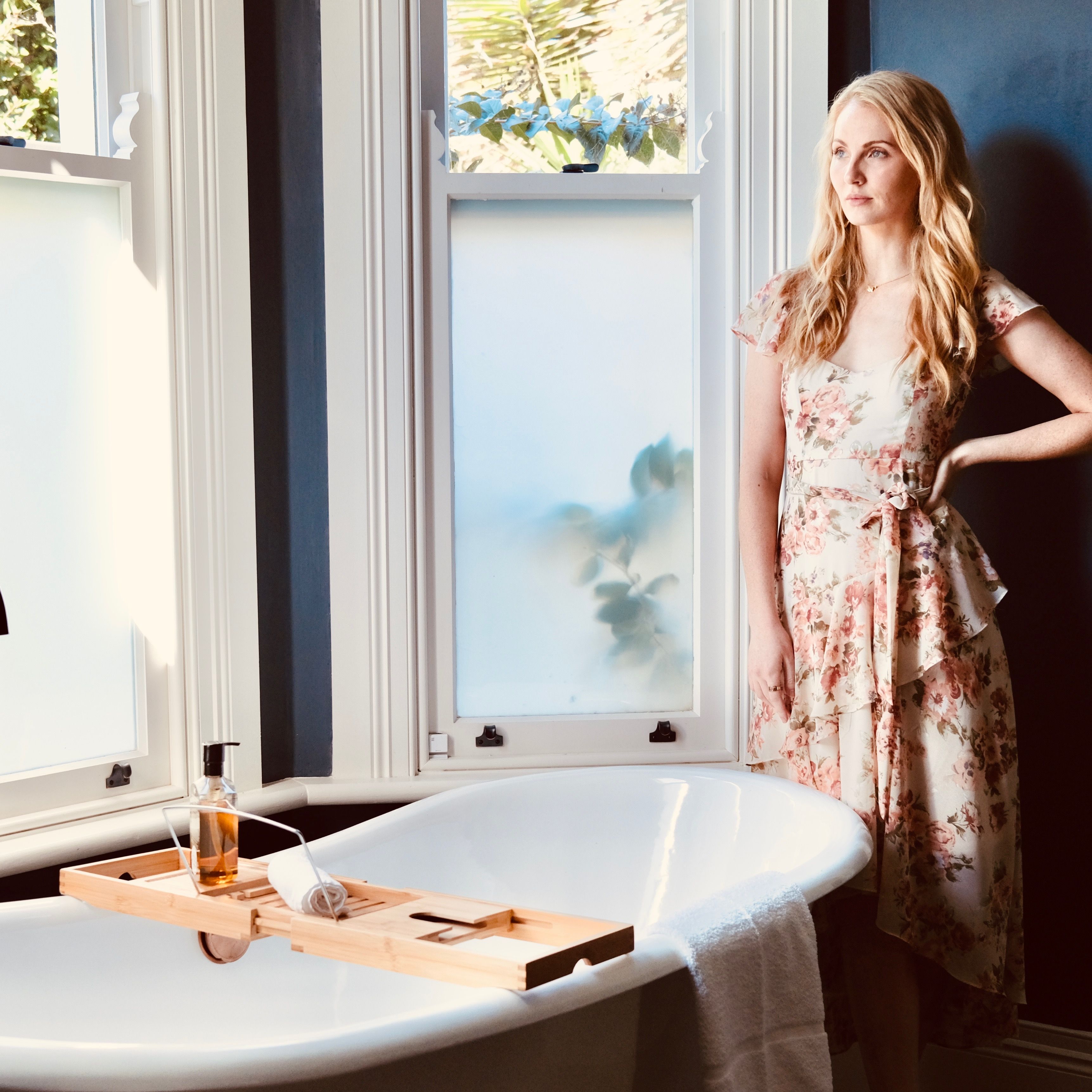 Woman in floral dress standing by bathtub in bright modern bathroom.