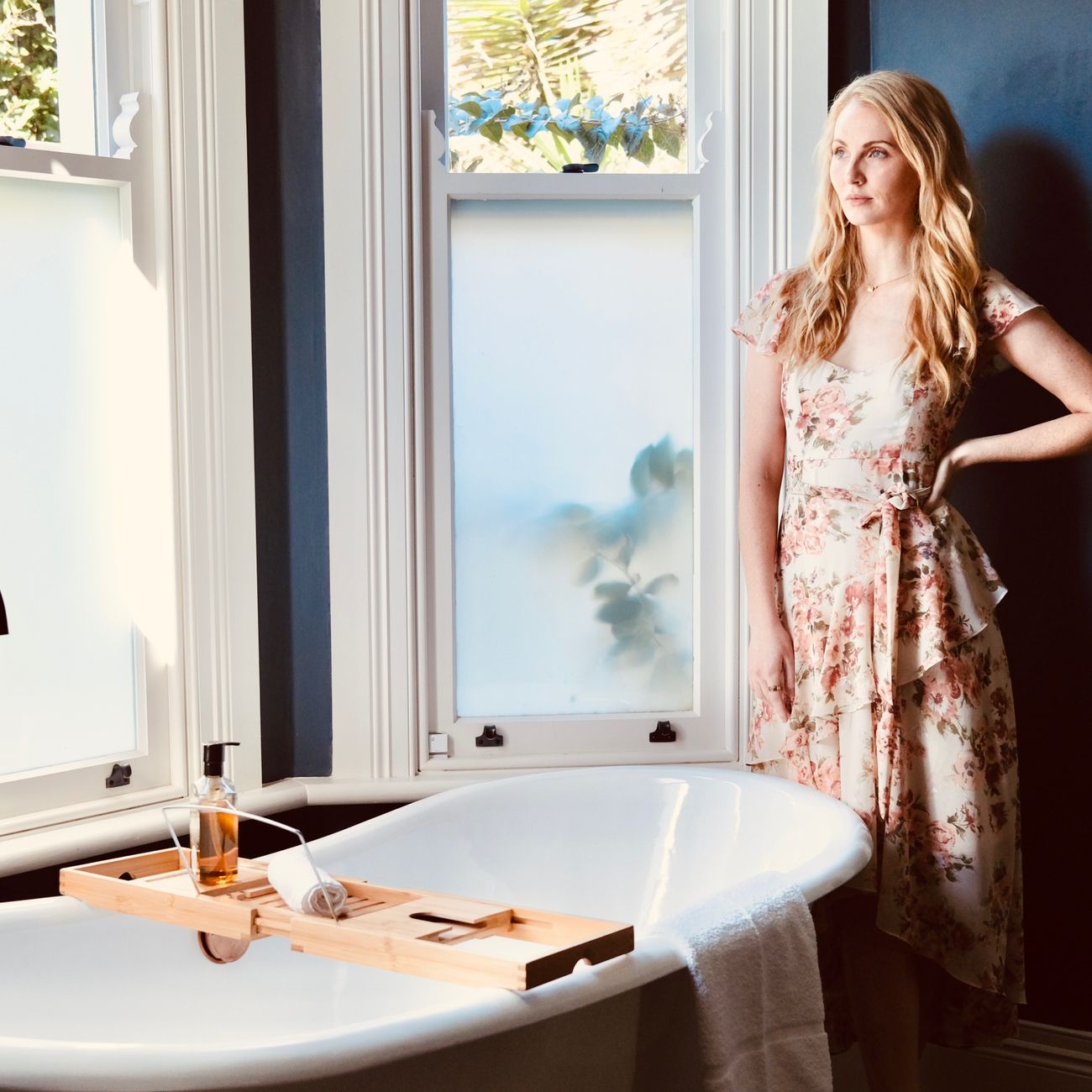 Woman in floral dress standing by bathtub in bright modern bathroom.
