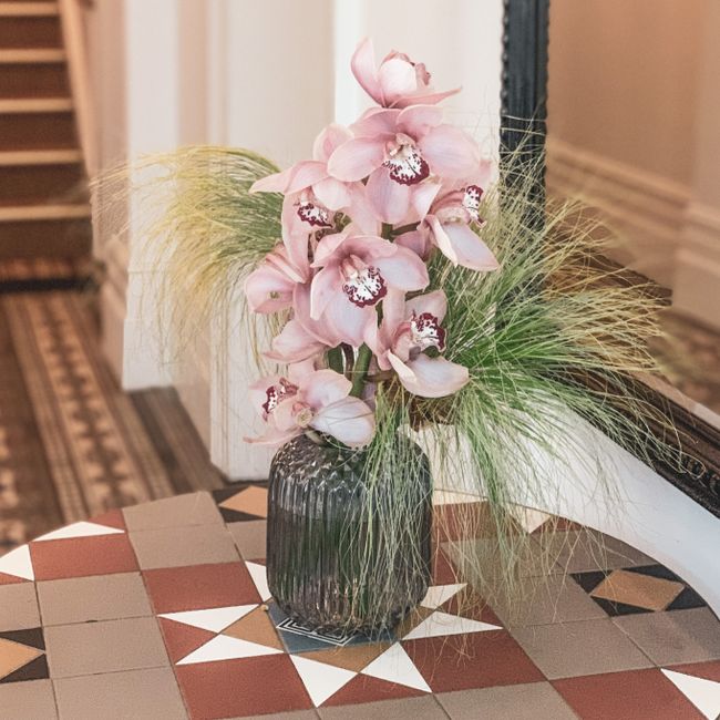 A stylish vase with pink orchids and decorative grass on a tiled table indoors.