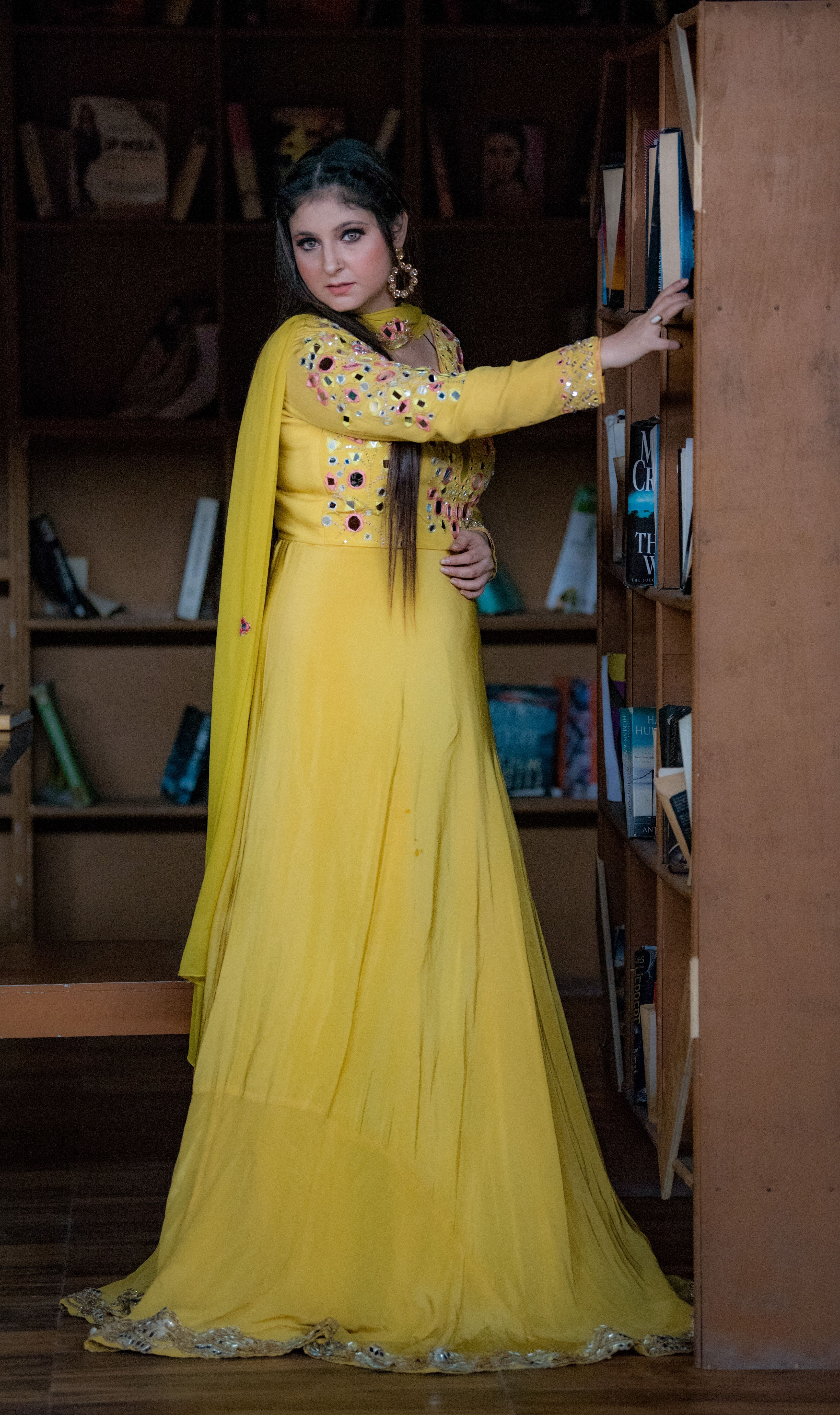 A young woman gracefully poses in a yellow traditional dress inside a library, exuding elegance.