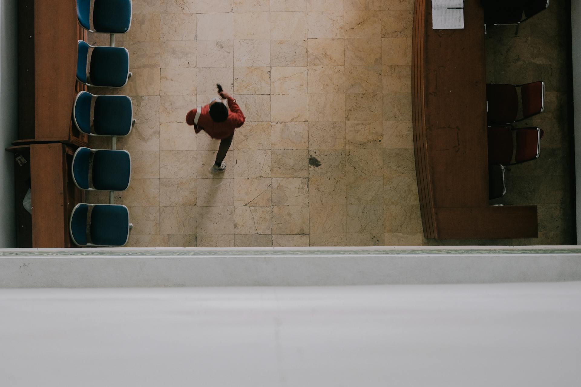 Aerial shot of a man walking in a tiled indoor corridor with chairs aligned on the side.