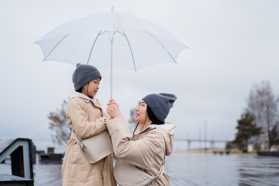 Mother and daughter in matching coats and beanies holding an umbrella, enjoying a rainy day outdoors.