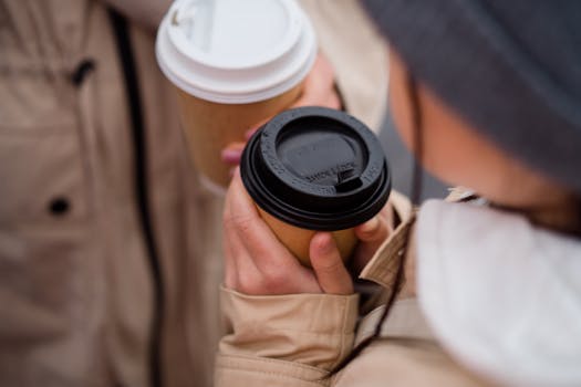 Close-up of two people holding takeaway coffee cups with lids, perfect for a warm, cozy day outdoors.