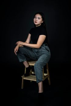 Chic portrait of a woman in denim overalls sitting on wooden chair against dark backdrop.