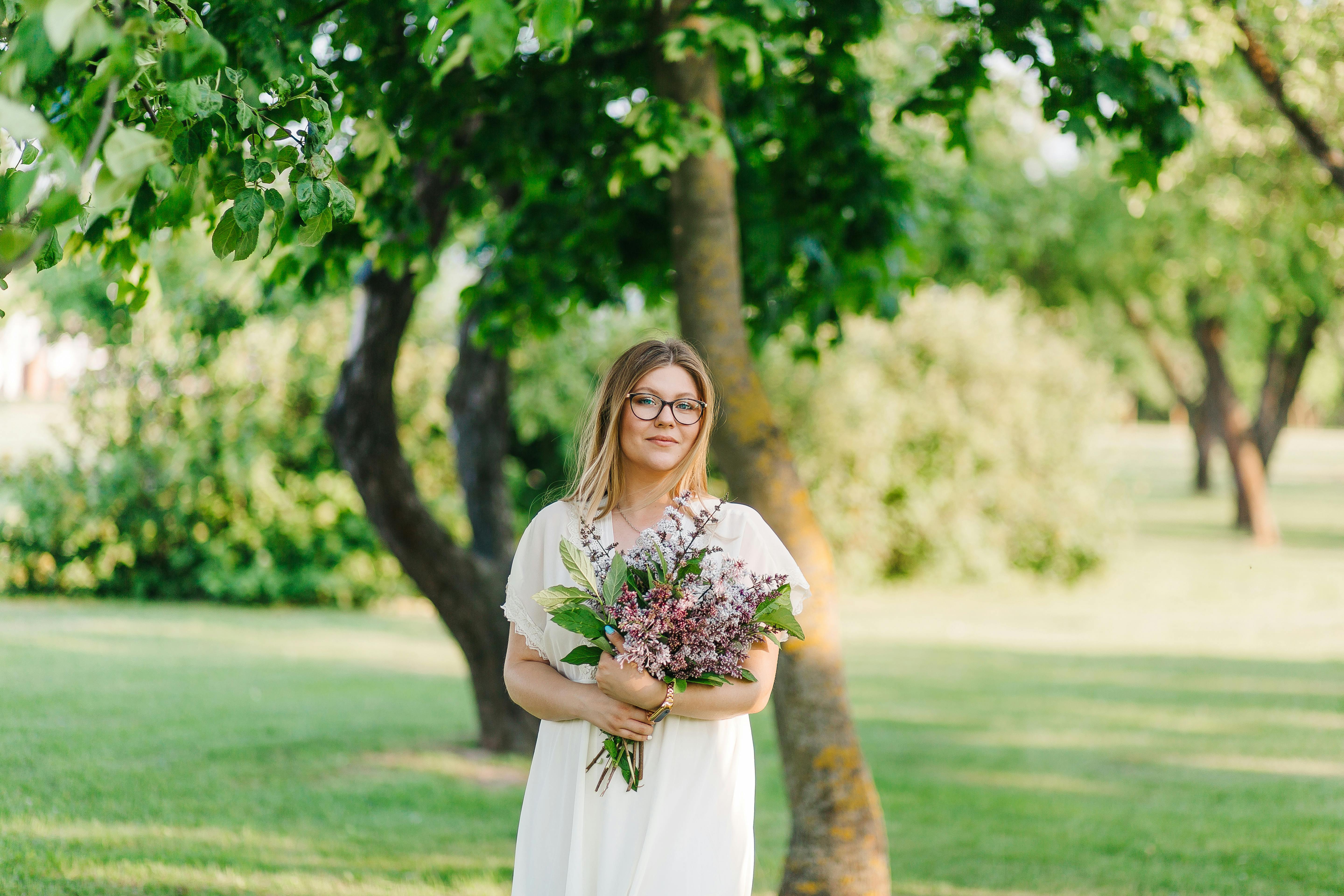Woman in white dress holds a bouquet of flowers in a sunny park setting.