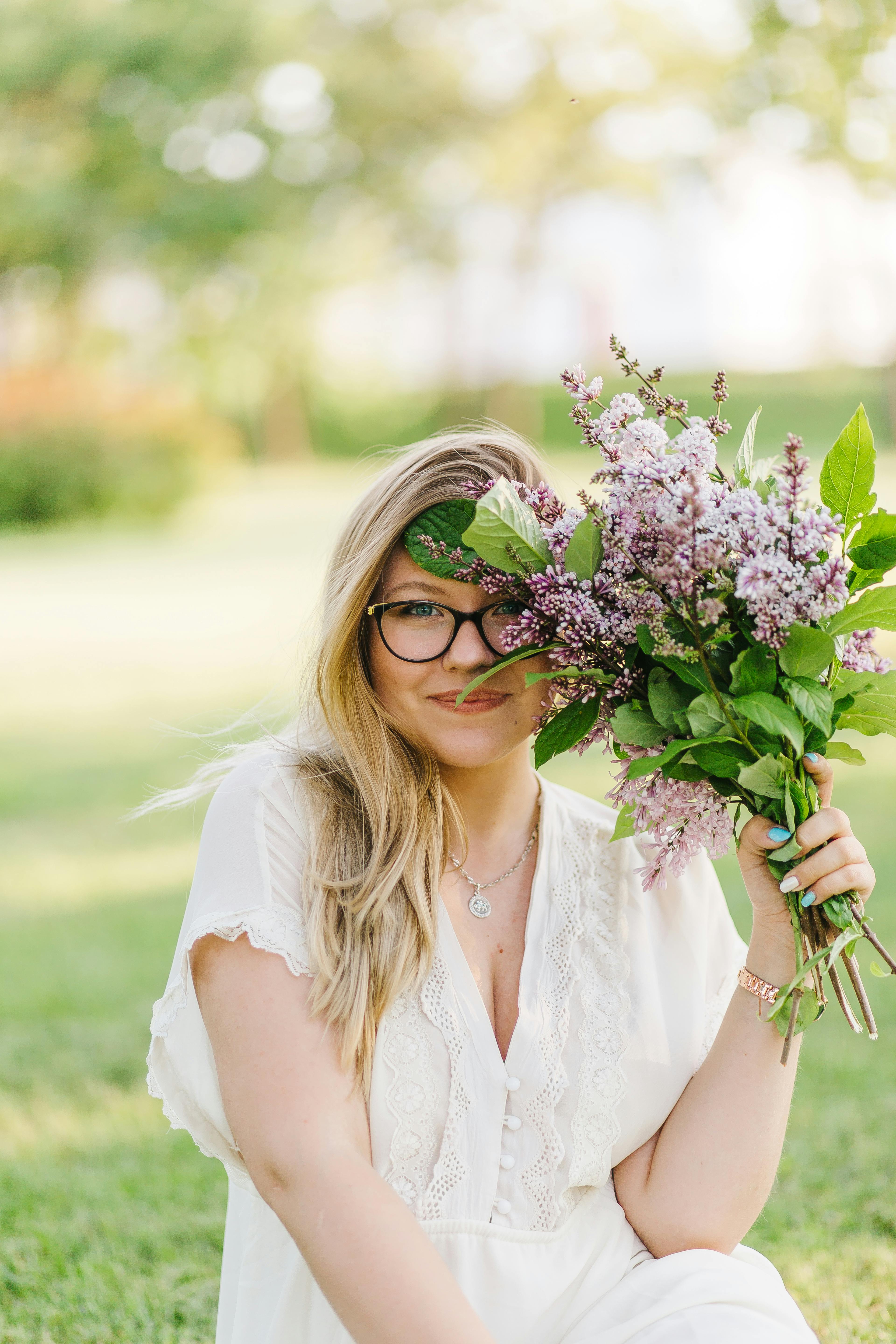 A cheerful woman in a white blouse sits outdoors, holding a vibrant lilac bouquet.