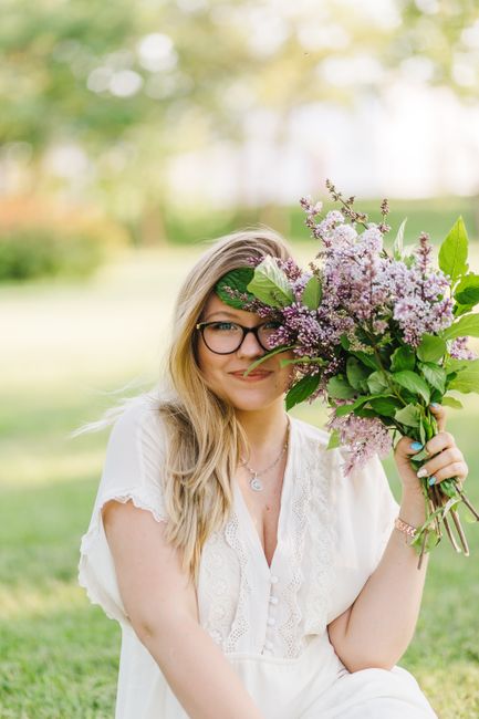 A cheerful woman in a white blouse sits outdoors, holding a vibrant lilac bouquet.