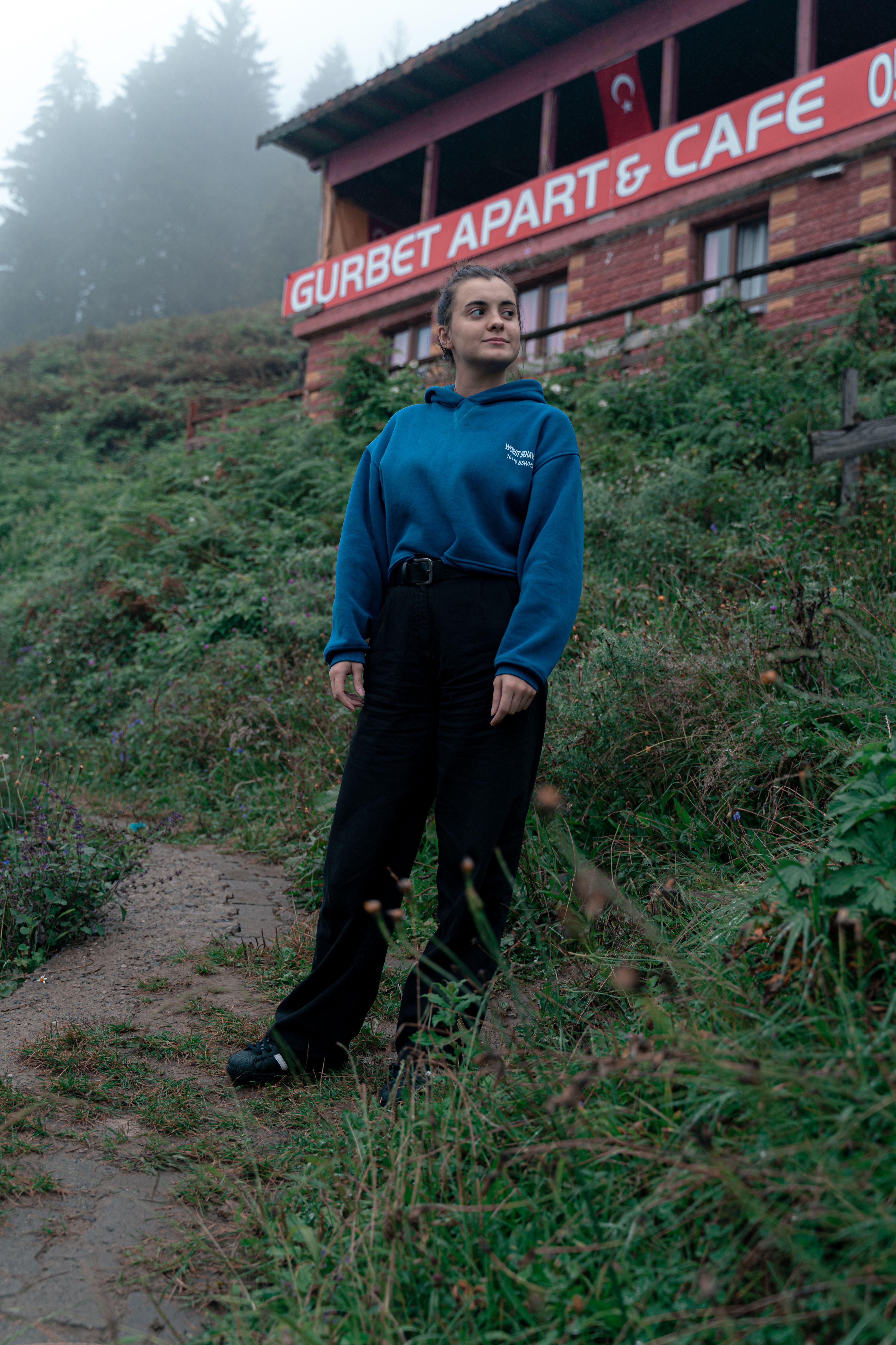 Woman in blue jacket standing by Gurbet Apart Cafe, surrounded by nature in Turkey.