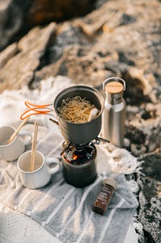 A camping setup featuring instant noodles cooking on a portable gas stove with cups and utensils.