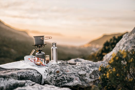 A scenic outdoor camping setup with cooking gear on rocky terrain during sunrise in a mountain landscape.
