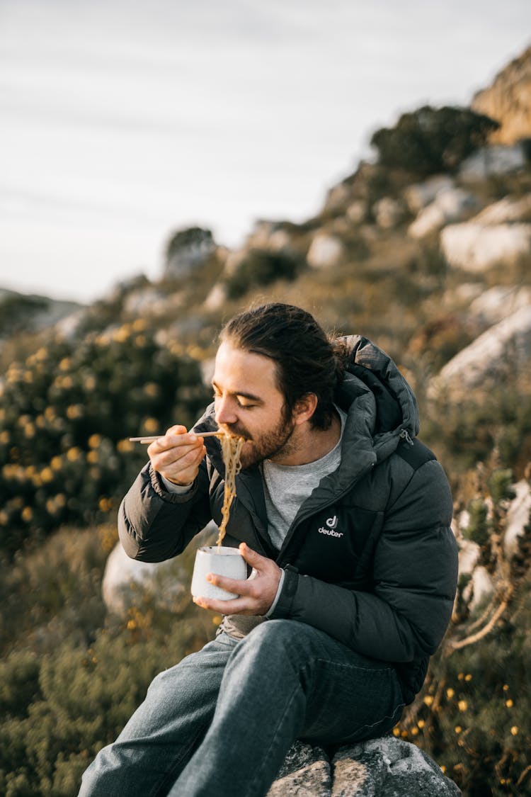 A Man In Black Jacket Sitting On Big Rock While Eating Noodles