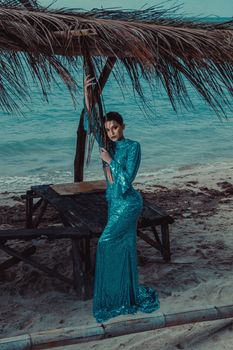 Stylish woman wearing a blue sequin gown poses by a rustic hut on a sandy beach.