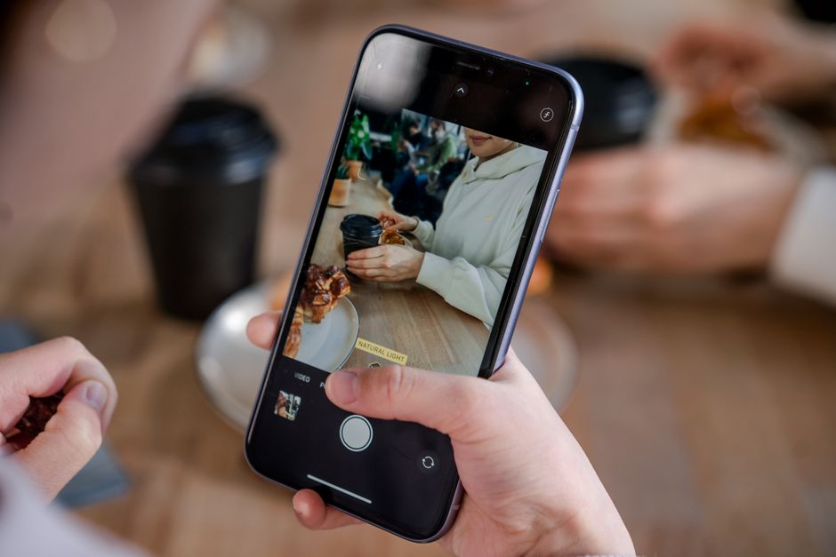 A person holding a smartphone capturing a cafe scene with a focus on coffee and pastry.