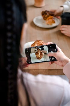 A woman photographing a croissant with her smartphone in a cozy cafe setting.