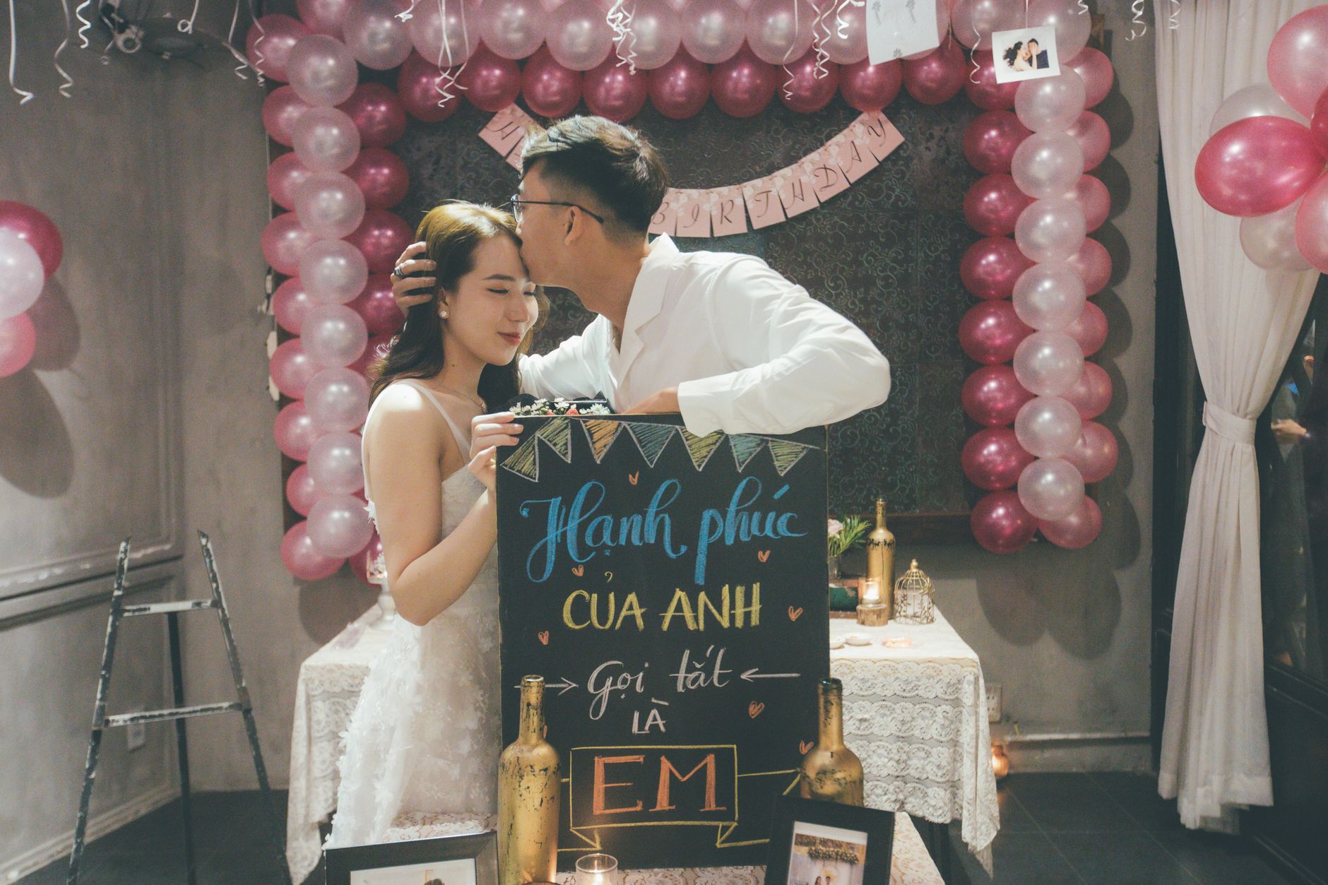 A couple shares a tender moment during a romantic celebration with balloons and a decorative sign.