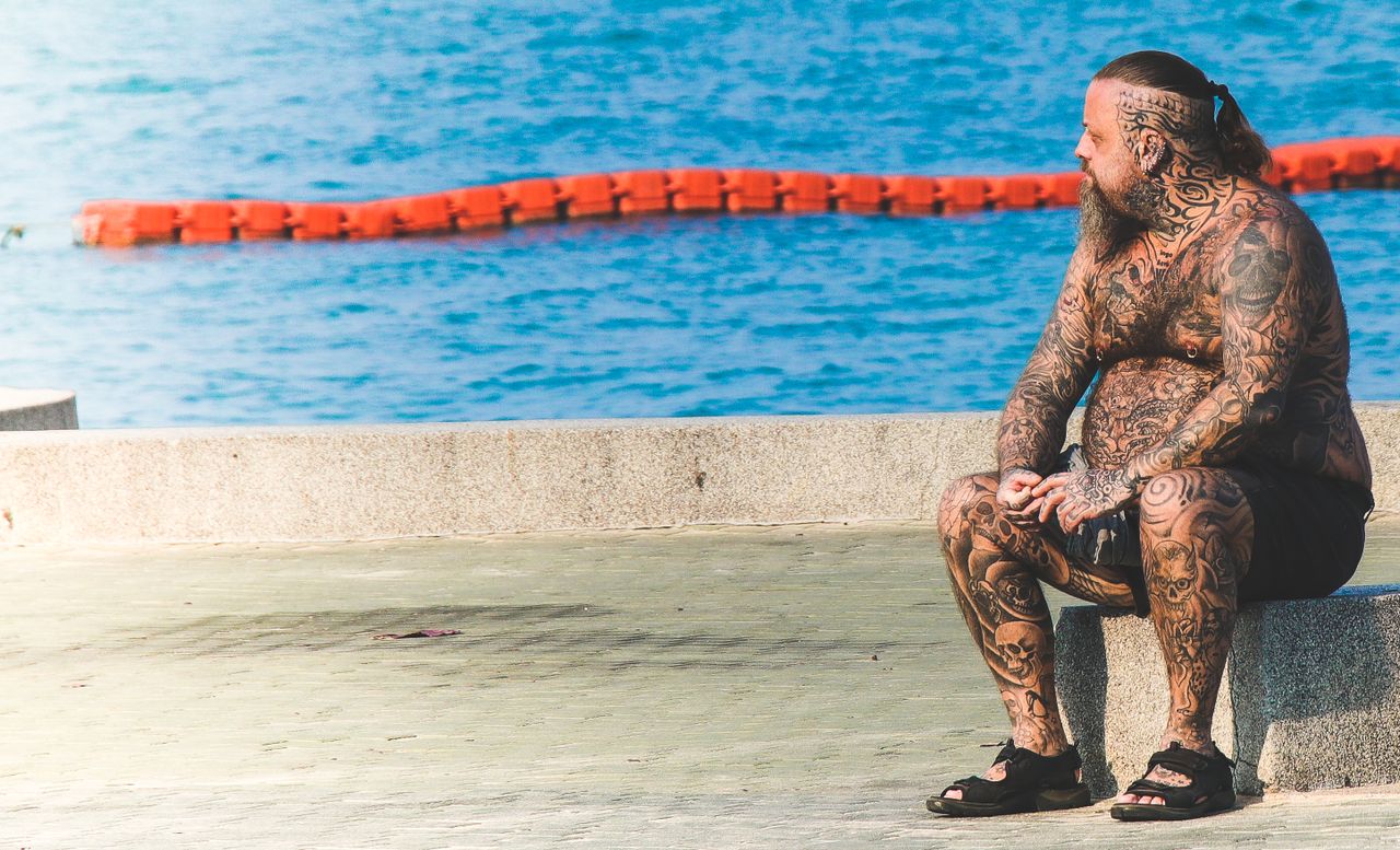 a heavily tattooed man sitting by the ocean enjoying a sunny day by th