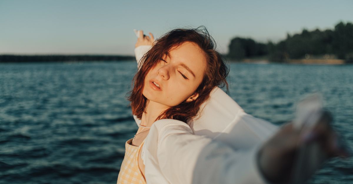 A peaceful woman basks in the summer sun with eyes closed and arms outstretched by a calm lake.