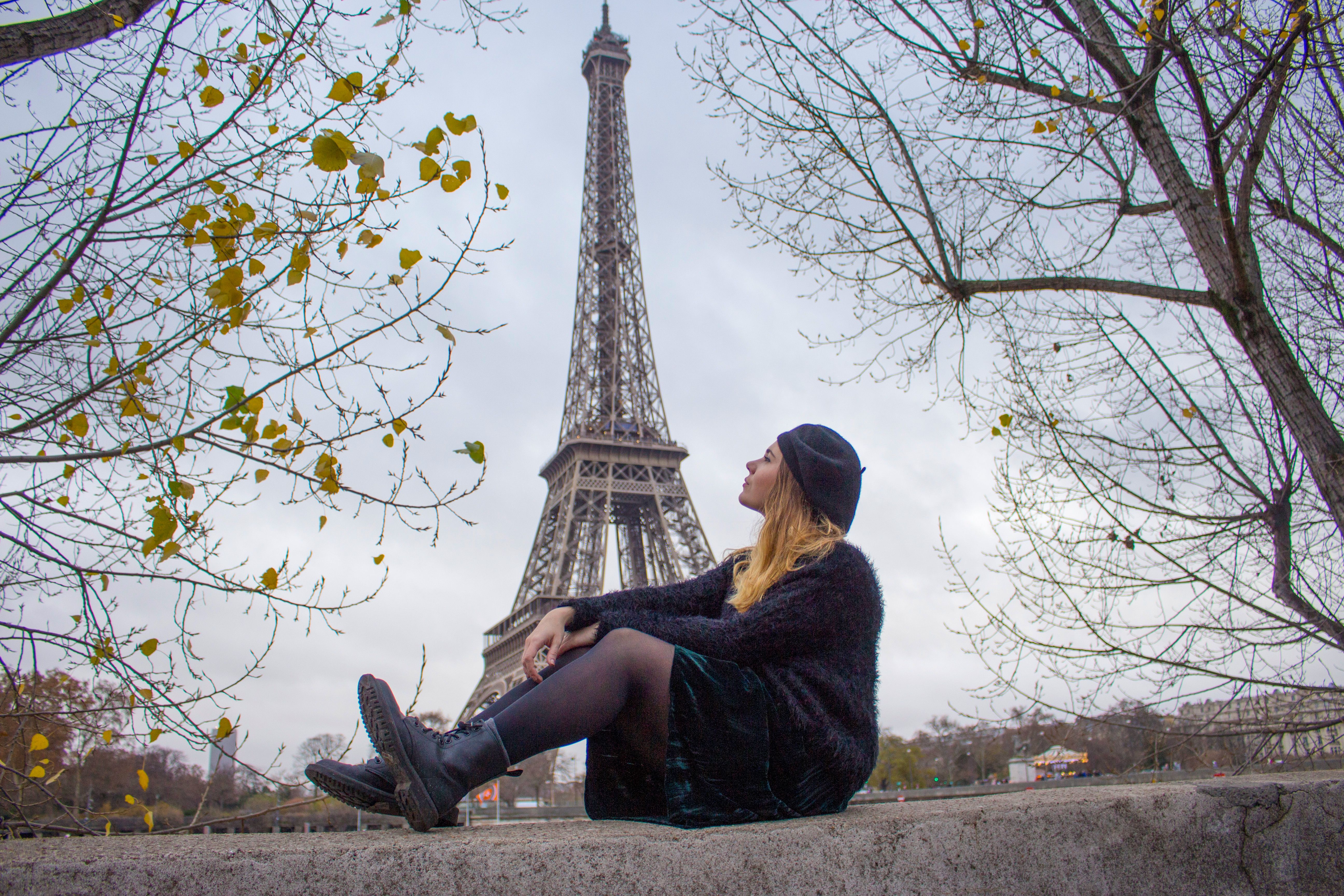 A woman in a stylish outfit sits contemplatively near the Eiffel Tower during fall in Paris.