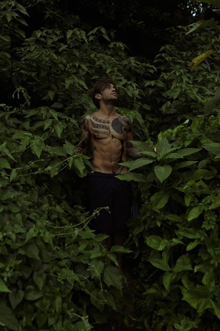 Muscular tattooed man amidst dense foliage in an outdoor forest photoshoot.