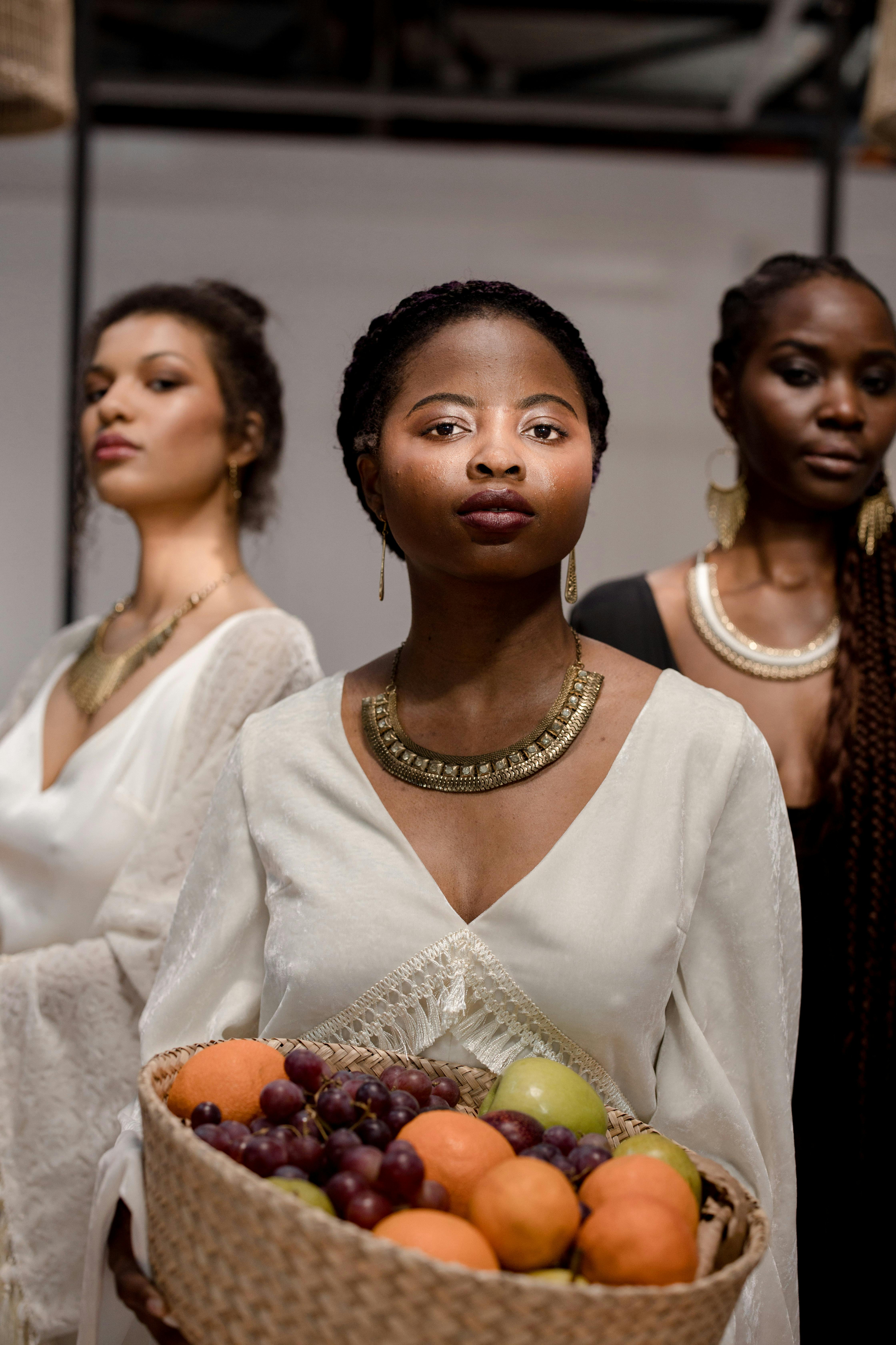 Three elegant women in stylish dresses posing with a fruit basket, showcasing fashion and diversity.