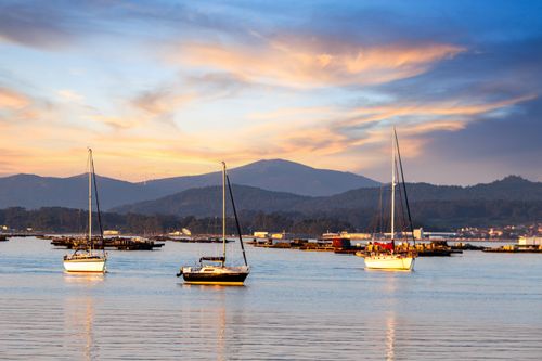 Free Scenic view of sailboats at sunset in El Grove, Spain, with mountains and a colorful sky. Stock Photo