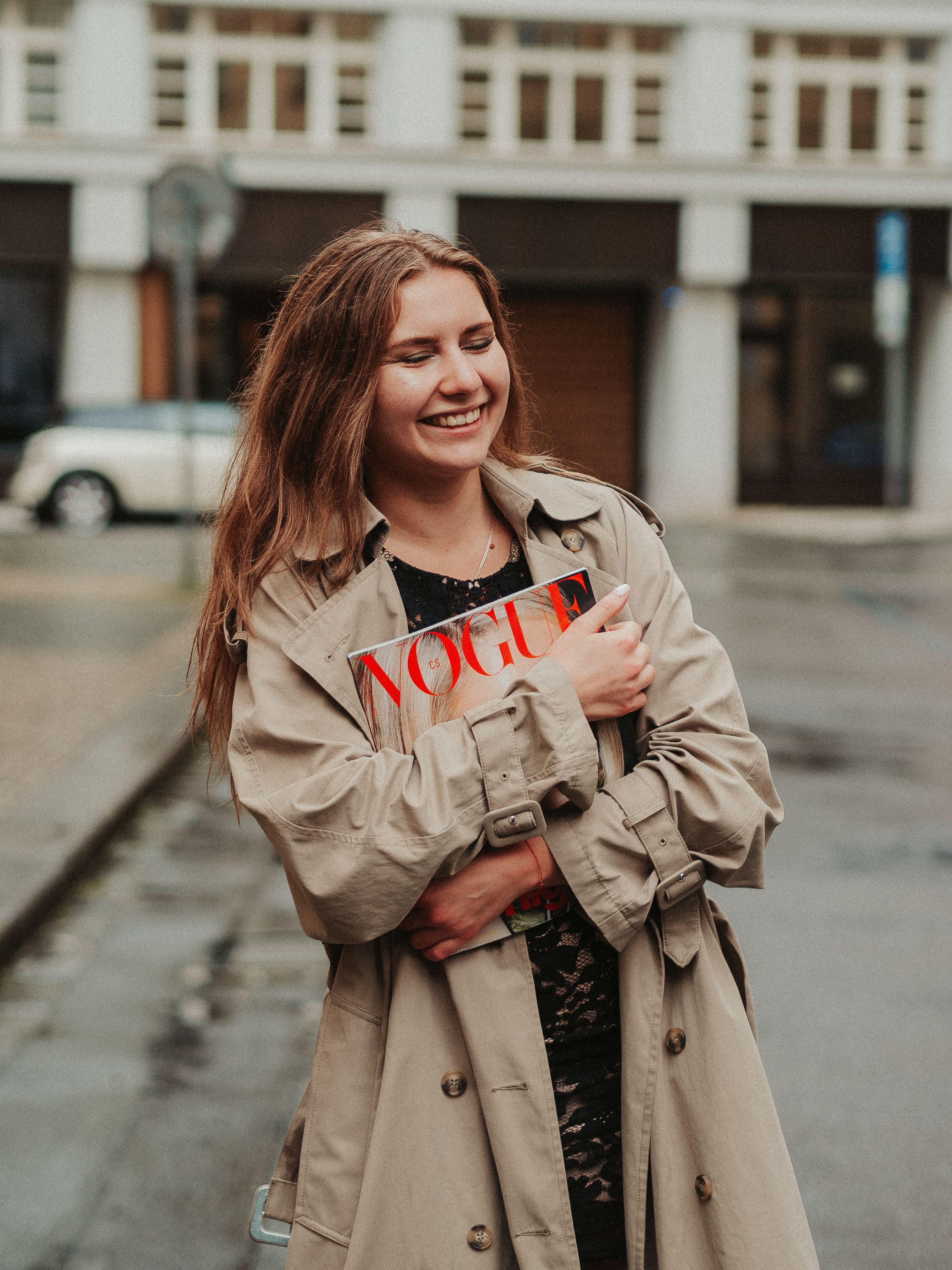 A cheerful woman stands outdoors holding a Vogue magazine, dressed in a trench coat.