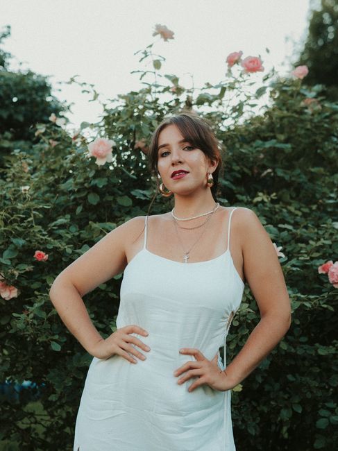 A woman in a white dress poses gracefully among blooming roses in a garden.