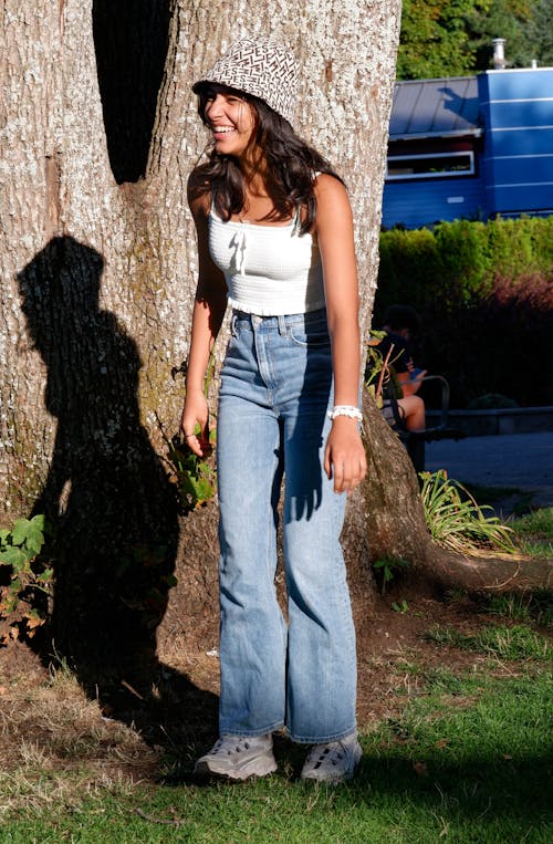 Woman in White Tank Top and Blue Denim Jeans Standing Beside Brown Tree