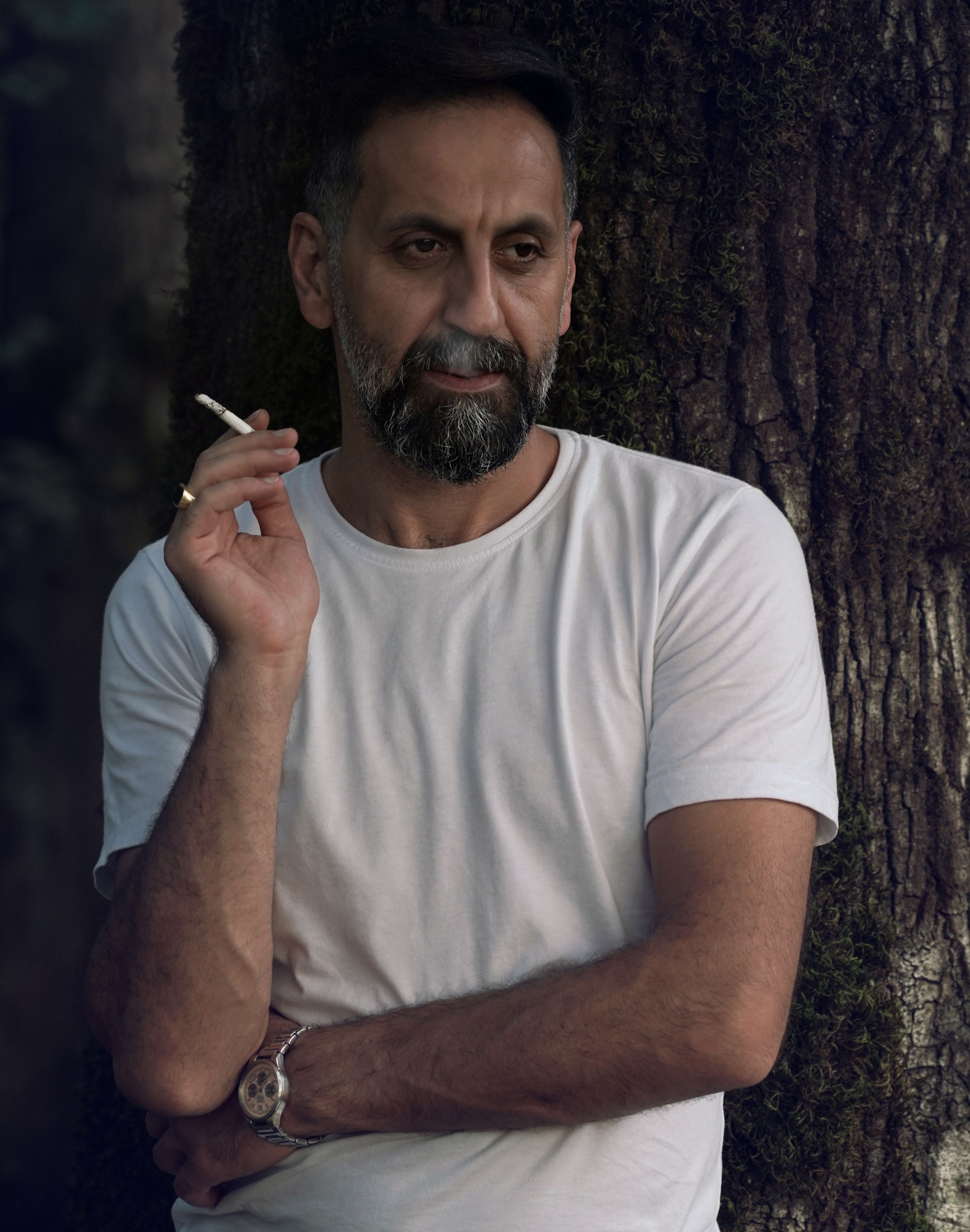 A thoughtful Middle Eastern man in a white shirt smokes outdoors by a tree.