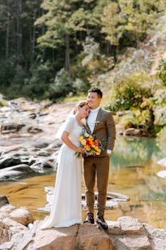 Newlyweds embrace beside a serene forest stream, holding a vibrant bouquet.