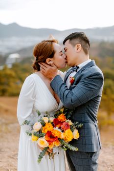 Couple kissing outdoors in wedding attire, holding a vibrant floral bouquet.
