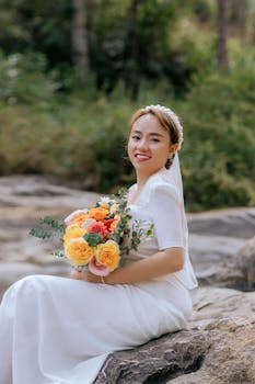 Smiling bride holding a vibrant bouquet, seated outdoors in a natural setting.
