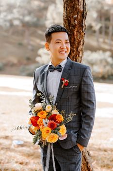 Asian groom smiling brightly while holding a colorful wedding bouquet outdoors under natural light.