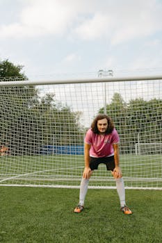 Soccer player in pink jersey stands confidently in goal, ready for action on an outdoor soccer field.