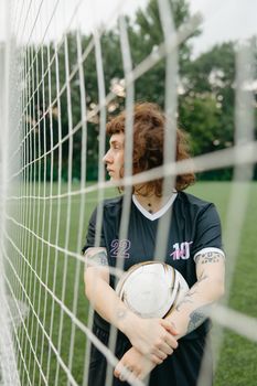 A female soccer player posing with a ball by the goal net on a green field.