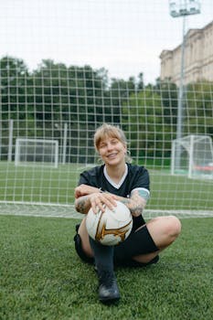 Caucasian woman soccer player in black uniform smiling while sitting with a soccer ball on a field.
