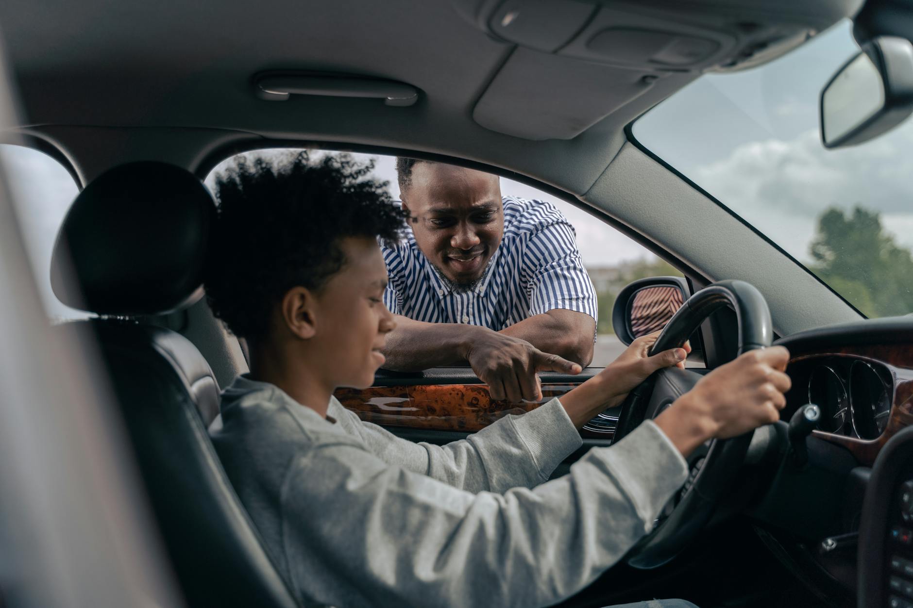 Young boy sitting at the wheel while father instructs from outside, learning to drive.