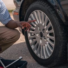 Man in a striped shirt crouching by a car's alloy wheel in daylight, pointing at the tyre