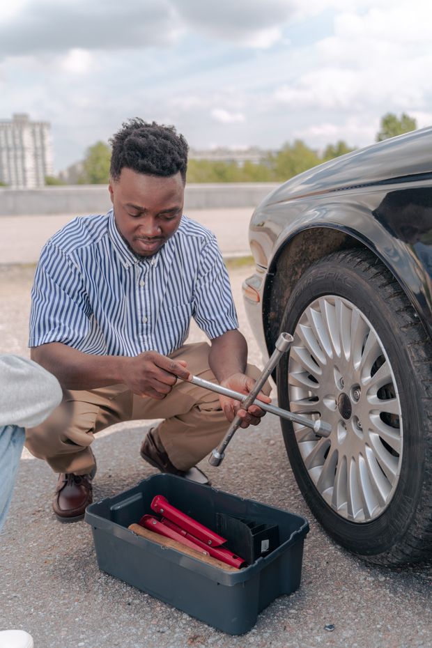 A man crouched beside a car on the road, turning a lug wrench on the front wheel