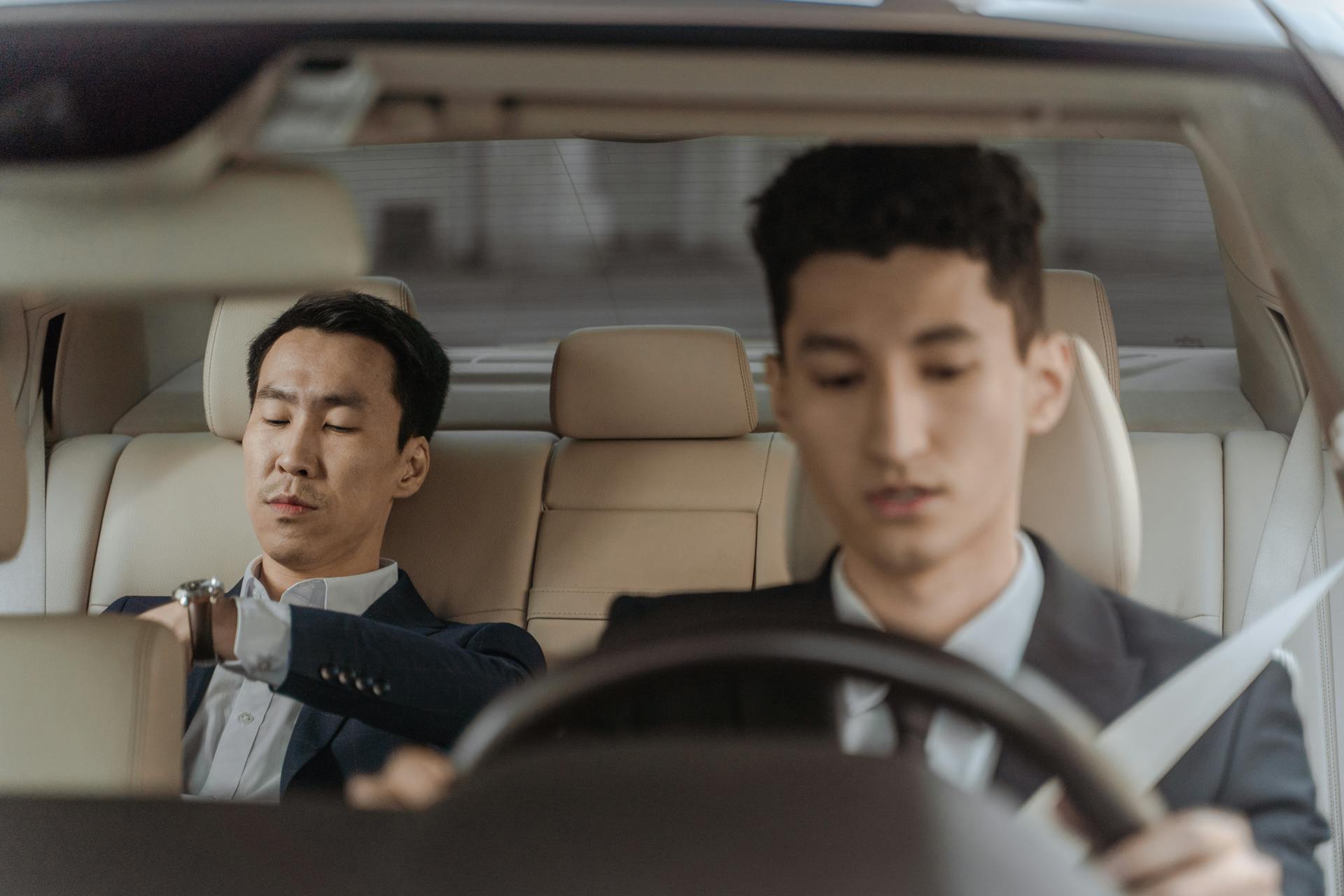 Two business professionals in suits inside a car, focused on driving and checking time.